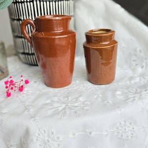 Puede incluir: Dos pequeños tarros de cerámica marrón, uno con asa, sobre un mantel de encaje blanco.