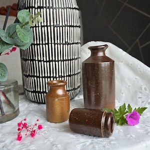 Puede incluir: Tres jarrones de cerámica de diferentes tamaños y estilos. El jarrón más grande es blanco y negro con un patrón geométrico. Los dos jarrones más pequeños son marrones y tienen un aspecto rústico. Los jarrones están sobre un mantel blanco con un patrón floral.