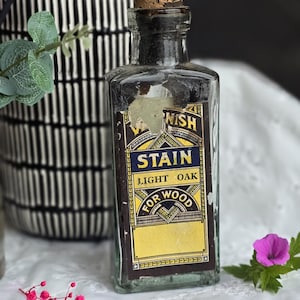 May include: A vintage glass bottle with a cork stopper and a label that reads "Varnish Stain Light Oak For Wood". The bottle is sitting on a white lace tablecloth with a small pink flower.