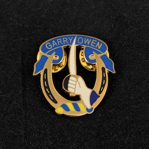 May include: Gold-colored pin with a blue and white horseshoe design. The horseshoe is surrounded by a blue ribbon with the text "GARRY OWEN" in white letters.