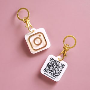 Peut inclure: Deux porte-clés blancs avec des accents dorés. Un porte-clés présente un logo Instagram doré et l'autre porte-clés présente un code QR doré avec le texte "@nattu.artwork".