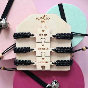 May include: Three black friendship bracelets with wooden puzzle pieces that say "Big Sis", "Middle Sis", and "Little Sis". The bracelets are displayed on a wooden stand with the word "natu" engraved on it.