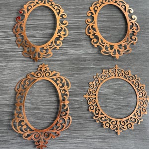 May include: Four copper-colored, ornate frames in oval and circular shapes. Each frame features intricate scrollwork and decorative elements. The frames are arranged on a gray wooden surface, showcasing their detailed designs.