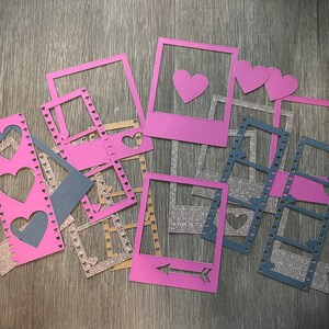 May include: A set of pink and grey paper frames with heart cut-outs. The frames are in the shape of film strips and instant photos.