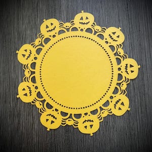 May include: Yellow paper doily with a lace design and a circle in the center. The doily is decorated with cut-out jack-o'-lantern faces around the edge.