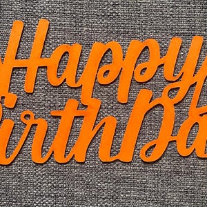 May include: A bright orange cutout sign featuring the words Happy Birthday in a cursive script font. The design includes two heart shapes integrated into the lettering, one positioned above the letter H and one above the letter y at the end.