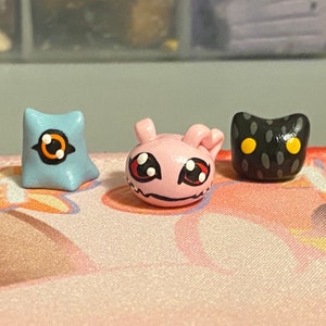 Op de afbeelding: Drie kleine kleifiguurtjes, elk met een ander ontwerp. De eerste is een lichtblauw figuurtje met een zwart en oranje oog. De tweede is een roze figuurtje met rode ogen en een zwarte mond. De derde is een zwart figuurtje met gele ogen en een getextureerd oppervlak.