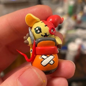May include: A hand-painted figurine of a cartoon character with yellow and red coloring. The character has a knife, a red horn, and an orange barrel-shaped body with white cross markings. The figure has a cute, friendly expression.