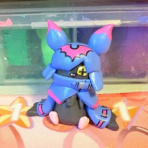 May include: A handmade cartoon character figurine, predominantly blue with pink detailing. The figure has large ears, a bat-shaped emblem, and yellow eyes. The character is posed with its arms crossed and small pink feet.