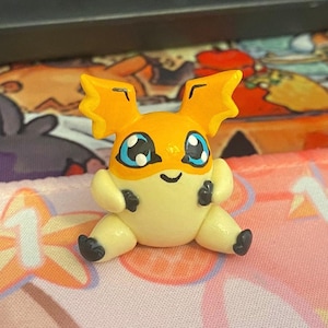 May include: A small, orange and yellow figurine of a cartoon character with large blue eyes and black accents. The figurine has large ears and is sitting with its legs out. The background is a patterned fabric.