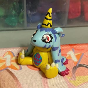 May include: A small, hand-sculpted figurine of a creature with a light blue head featuring dark blue stripes, a yellow horn with black markings, and large red eyes. The body is yellow with a blue and pink oval emblem on the chest and pink claws.