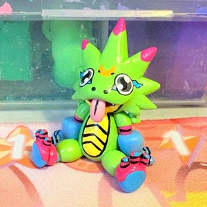 以下が含まれることがあります： 手作りの、緑色の漫画のような生き物のフィギュア。とげのある頭、ピンクのアクセント、長いピンクの舌があります。大きな目、青とピンクの足、黄色と黒の縞模様の腹を持っています。フィギュアは座っています。