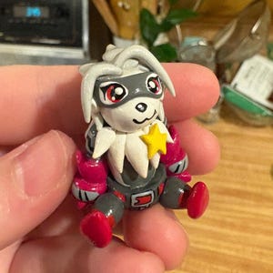 May include: A handmade figurine of a cartoon character with a white face, gray mask, and red eyes. The figure has a yellow star on its chest, pink gloves, and red boots. The figure is sitting.