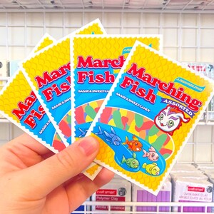 Pu&ograve; includere: Una mano tiene una pila di quattro pacchetti gialli e blu di caramelle "Marching Fish". I pacchetti presentano un pesce dei cartoni animati con una coda arcobaleno e il testo "Sassy & Sweet Candy".