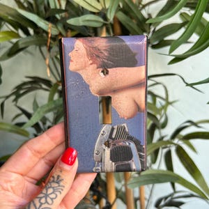 May include: A rectangular light switch cover plate featuring a printed graphic of a person with dark hair and a motorcycle engine. The plate has a central vertical rectangular cutout and two small circular screw holes for mounting purposes.