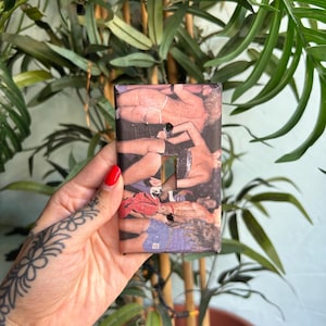 May include: A rectangular light switch wall plate featuring a collage of photographic imagery depicting multiple figures in various poses. The surface is printed with a multi-coloured graphic design and includes two pre-cut openings for switch toggles.