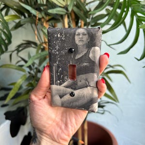 May include: A rectangular light switch cover plate featuring a black and white photographic print of a person sitting in a chair. The cover includes a central toggle switch cutout and two black screws positioned at the top and bottom for mounting.