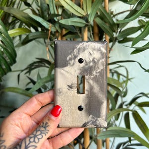 May include: A rectangular light switch cover plate featuring a black and white photographic print of a person with wavy hair. The cover plate has a central vertical toggle switch cutout and two small black screws positioned above and below the opening.