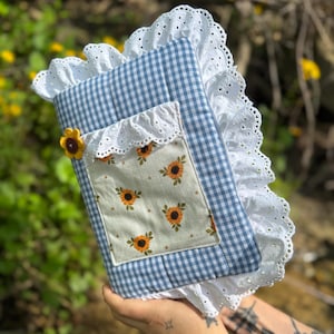 May include: A blue and white gingham fabric pouch with a white lace trim. The pouch has a pocket with a sunflower print and a small sunflower button.