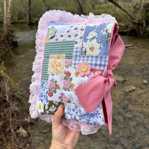 May include: A handmade, patchwork journal with a pink bow closure. The cover features floral and patterned fabric squares, accented with pink lace trim and crocheted flower embellishments. The journal is held up against a natural background.