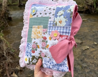 May include: A handmade, patchwork journal with a pink bow closure. The cover features floral and patterned fabric squares, accented with pink lace trim and crocheted flower embellishments. The journal is held up against a natural background.