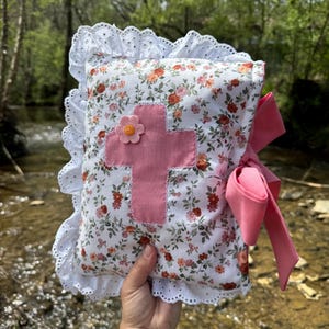 May include: A white pillow with a pink floral pattern and a pink cross with a flower in the center. The pillow has white lace trim and a pink ribbon.