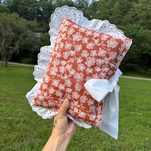 May include: A floral print pillow with a white eyelet lace trim. The pillow has a rust-colored background with white floral patterns. A white fabric is attached to the pillow.
