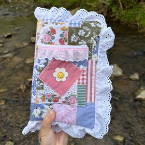 May include: A handmade journal with a patchwork design. The cover features floral patterns, pink lace, and a white daisy with a yellow button center. The edges are trimmed with white eyelet lace. The journal is held against a natural background.