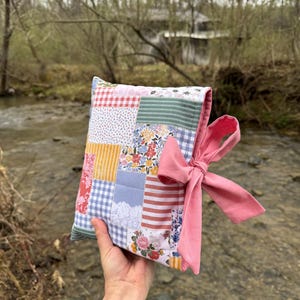 May include: A patchwork fabric pouch with a pink ribbon tie. The pouch is made of various floral and geometric patterns in shades of pink, blue, yellow, and green.