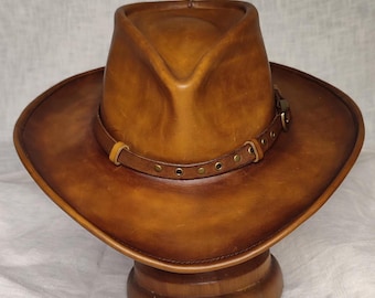 Könnte beinhalten: Brauner Leder-Cowboyhut mit breiter Krempe und Lederband mit Messingnieten. Der Hut wird auf einem hölzernen Hutständer präsentiert. Klassischer Western-Stil.
