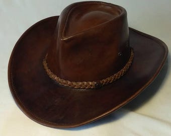 Könnte beinhalten: Ein brauner Cowboyhut aus Leder mit einem geflochtenen Band um den Kronenansatz. Der Hut hat eine breite Krempe und eine abgerundete Krone. Das Leder scheint abgenutzt zu sein, was ihm einen Vintage-Look verleiht. Der Hut ist ein klassischer Western-Stil.