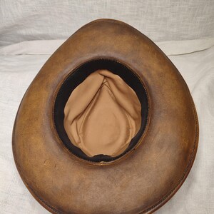 K&ouml;nnte beinhalten: Ein brauner Leder-Cowboyhut, von innen betrachtet, mit beigefarbenem Innenfutter und einem schwarzen Band. Der Hut hat eine breite, gebogene Krempe mit sichtbaren N&auml;hten. Das Design erinnert an den klassischen Western-Stil.