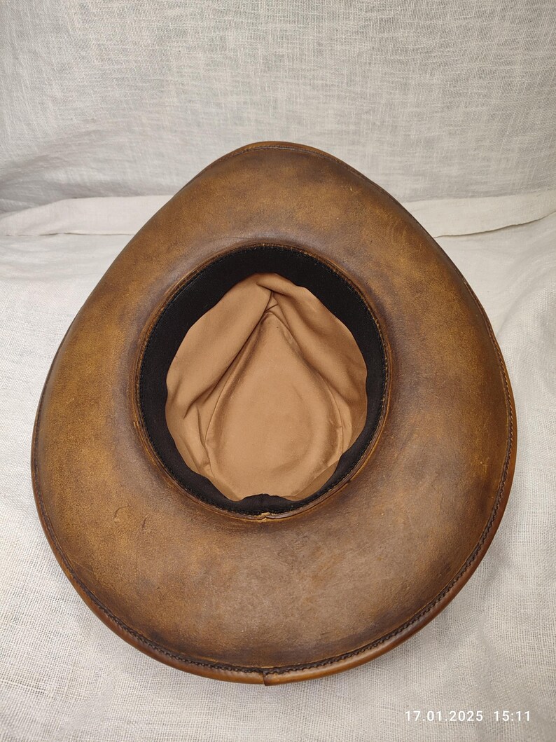 K&ouml;nnte beinhalten: Ein brauner Leder-Cowboyhut, von innen betrachtet, mit beigefarbenem Innenfutter und einem schwarzen Band. Der Hut hat eine breite, gebogene Krempe mit sichtbaren N&auml;hten. Das Design erinnert an den klassischen Western-Stil.