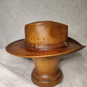 K&ouml;nnte beinhalten: Brauner Leder-Cowboyhut mit breiter Krempe und Lederband mit Messingnieten. Der Hut wird auf einem Holzst&auml;nder pr&auml;sentiert. Die Farbe des Hutes ist ein warmes, mittleres Braun, und er scheint aus hochwertigem Leder zu sein.