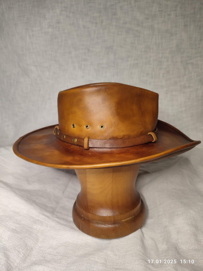 K&ouml;nnte beinhalten: Brauner Leder-Cowboyhut mit breiter Krempe und Lederband mit Messingnieten. Der Hut wird auf einem Holzst&auml;nder pr&auml;sentiert. Die Farbe des Hutes ist ein warmes, mittleres Braun, und er scheint aus hochwertigem Leder zu sein.