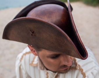 Könnte beinhalten: Ein brauner Leder-Tricorn-Hut mit breiter Krempe und einem gestickten X auf der Krone. Der Hut wird von einer Person mit einem weißen Hemd getragen.