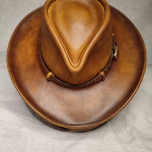 K&ouml;nnte beinhalten: Brauner Cowboyhut aus Leder mit breiter Krempe und spitz zulaufender Krone. Der Hut hat ein Lederband mit einer Messingschnalle. Ein klassischer Westernhut.