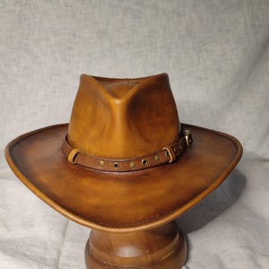 K&ouml;nnte beinhalten: Ein brauner Cowboyhut aus Leder mit breiter Krempe und Lederband, das mit Messingnieten befestigt ist. Der Hut wird auf einem h&ouml;lzernen Hutst&auml;nder pr&auml;sentiert. Der Hut hat einen klassischen Western-Stil.
