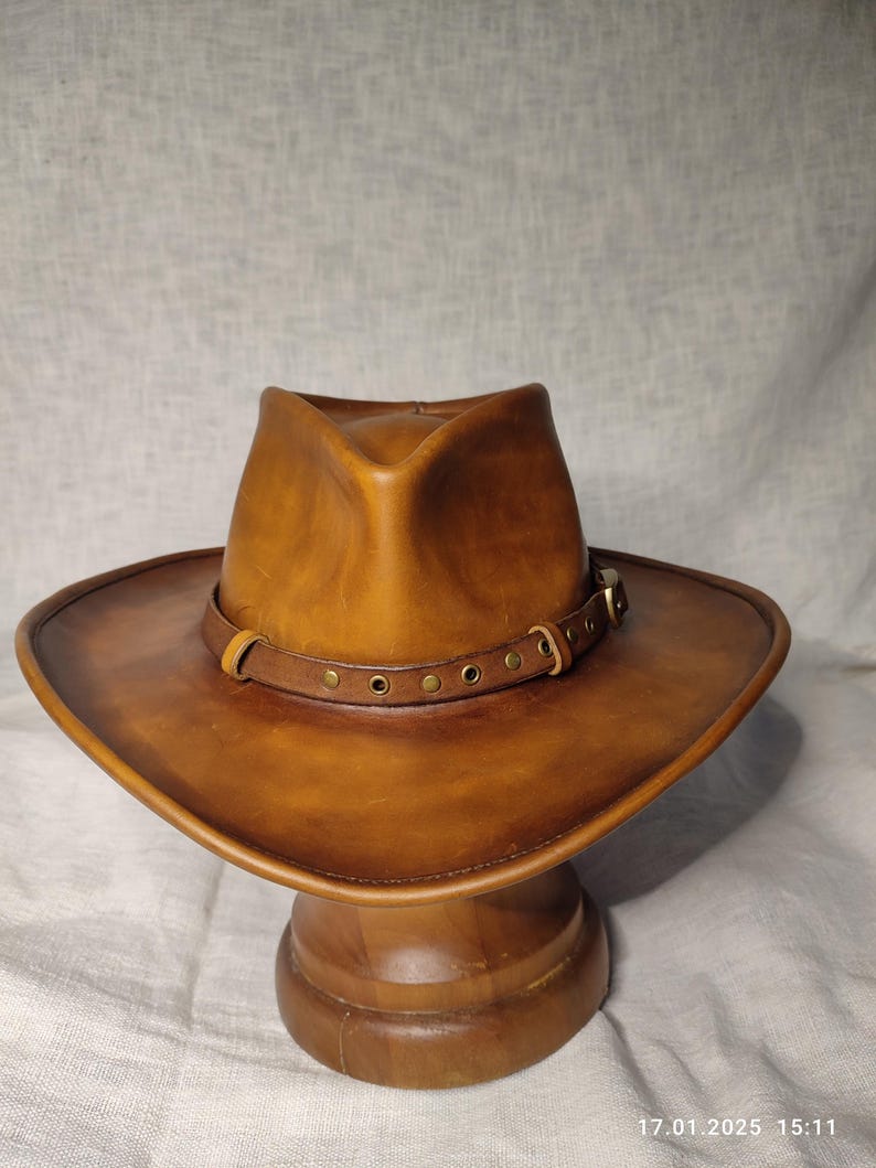 K&ouml;nnte beinhalten: Ein brauner Cowboyhut aus Leder mit breiter Krempe und Lederband, das mit Messingnieten befestigt ist. Der Hut wird auf einem h&ouml;lzernen Hutst&auml;nder pr&auml;sentiert. Der Hut hat einen klassischen Western-Stil.