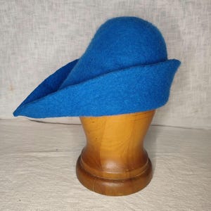 May include: A vibrant blue hat with a distinctive brim, displayed on a wooden hat stand. The hat's color is a bright, solid blue, and the brim extends outward. The hat is made of a soft, felt-like material.