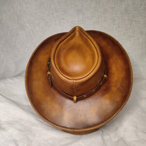 K&ouml;nnte beinhalten: Ein brauner Cowboyhut aus Leder mit runder Krone und breiter Krempe. Der Hut hat ein dekoratives Band mit Nieten und einen Lederriemen. Der Hut hat eine warme, mittelbraune Farbe und scheint aus hochwertigem Leder zu sein.