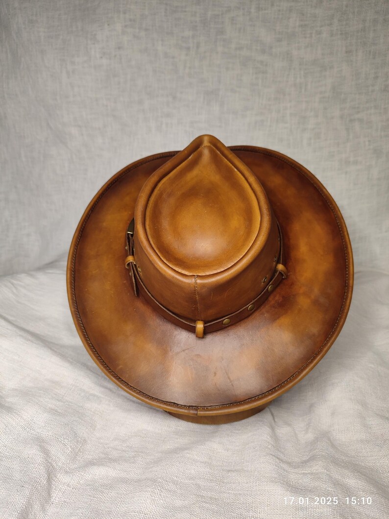 K&ouml;nnte beinhalten: Ein brauner Cowboyhut aus Leder mit runder Krone und breiter Krempe. Der Hut hat ein dekoratives Band mit Nieten und einen Lederriemen. Der Hut hat eine warme, mittelbraune Farbe und scheint aus hochwertigem Leder zu sein.