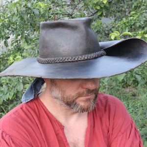 Könnte beinhalten: Ein dunkelbrauner Lederhut mit breiter Krempe und geflochtenem Band. Der Hut hat eine leicht gebogene Krone und ein Detail aus Denim-Stoff. Er ist für Sonnenschutz und Outdoor-Aktivitäten konzipiert.