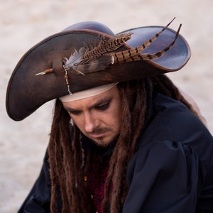 Könnte beinhalten: Ein brauner Piratenhut aus Leder mit breiter Krempe, verziert mit Federn und einer dekorativen Nadel. Der Hut wird von einer Person mit langen, braunen Dreadlocks und einem schwarzen Hemd getragen. Das Design des Hutes erinnert an Abenteuer und historische Kostüme.