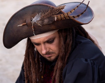 Könnte beinhalten: Ein brauner Piratenhut aus Leder mit breiter Krempe, verziert mit Federn und einer dekorativen Nadel. Der Hut wird von einer Person mit langen, braunen Dreadlocks und einem schwarzen Hemd getragen. Das Design des Hutes erinnert an Abenteuer und historische Kostüme.
