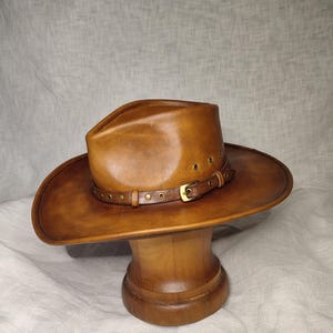 K&ouml;nnte beinhalten: Ein brauner Cowboyhut aus Leder mit breiter Krempe und Lederband mit Messingnieten. Der Hut wird auf einem h&ouml;lzernen Hutst&auml;nder pr&auml;sentiert. Der Hut hat einen klassischen Western-Stil.
