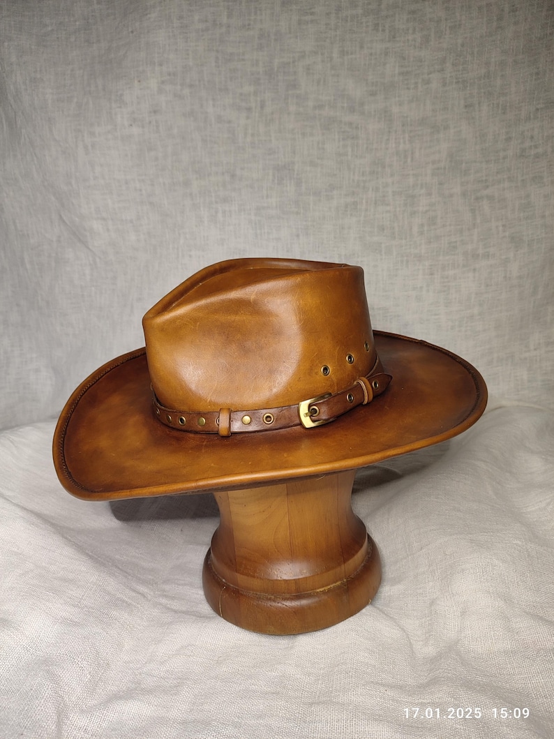 K&ouml;nnte beinhalten: Ein brauner Cowboyhut aus Leder mit breiter Krempe und Lederband mit Messingnieten. Der Hut wird auf einem h&ouml;lzernen Hutst&auml;nder pr&auml;sentiert. Der Hut hat einen klassischen Western-Stil.