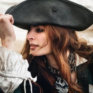 Könnte beinhalten: Eine Person trägt einen schwarzen Piratenhut mit einem Kreuzdetail. Die Person trägt auch eine schwarze Jacke, ein weißes Rüschenhemd und einen gemusterten Schal. Die Person hat lange, wellige, rotbraune Haare.
