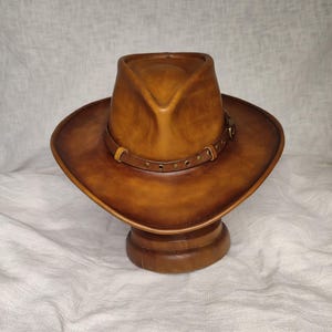 K&ouml;nnte beinhalten: Ein brauner Cowboyhut aus Leder mit breiter Krempe und einem dekorativen Band um den Kopf. Der Hut wird auf einem h&ouml;lzernen Hutst&auml;nder vor neutralem Hintergrund pr&auml;sentiert. Der Hut hat einen klassischen Western-Stil.