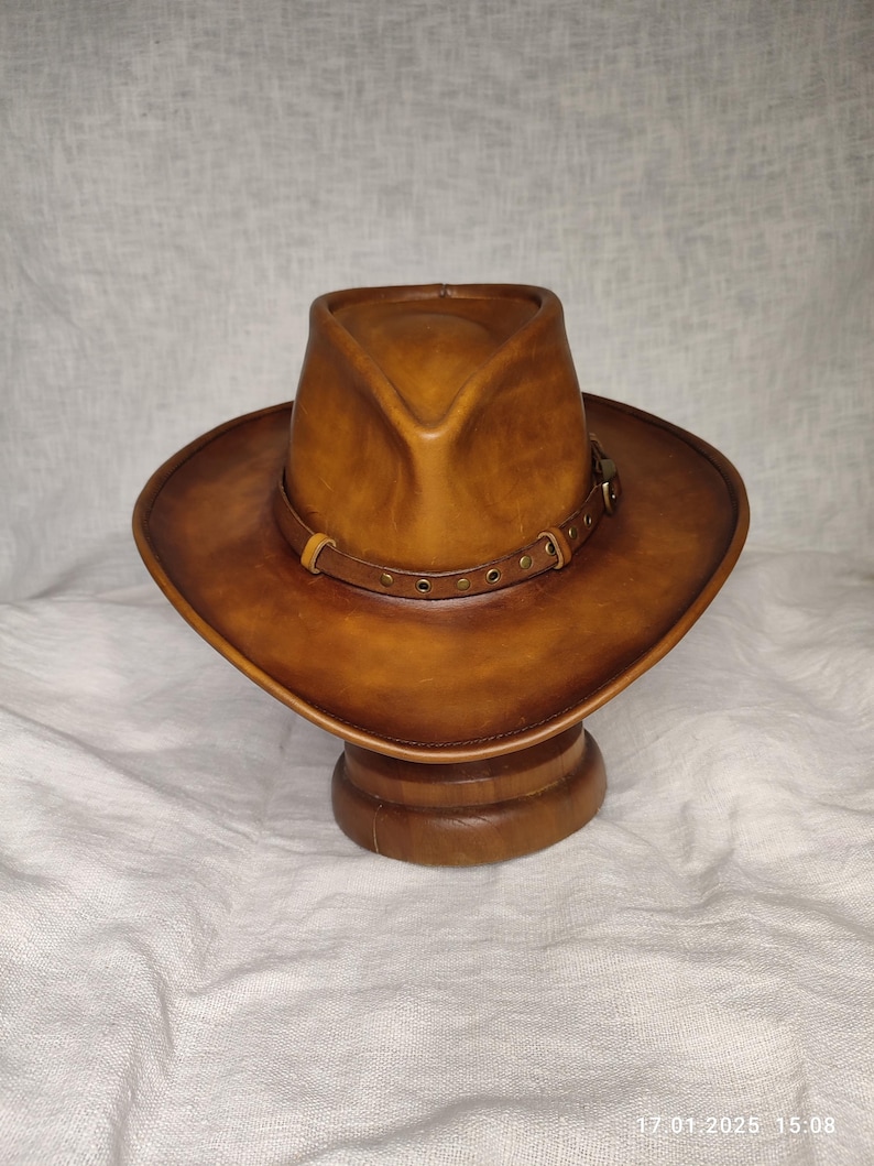 K&ouml;nnte beinhalten: Ein brauner Cowboyhut aus Leder mit breiter Krempe und einem dekorativen Band um den Kopf. Der Hut wird auf einem h&ouml;lzernen Hutst&auml;nder vor neutralem Hintergrund pr&auml;sentiert. Der Hut hat einen klassischen Western-Stil.